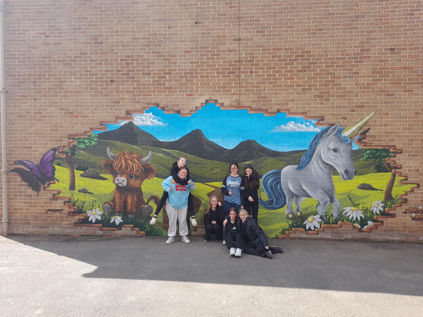 Seven members of the Blairgowrie High School Champs Board pose in front of their mural. The mural shows the wall breaking away to show a green, hilly landscape. A butterfly and unicorn are painted as emerging from it, while a young highland cow is painted to sit on the grass.